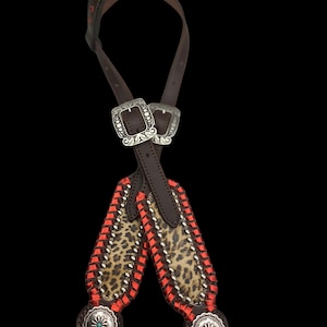 May include: A brown leather horse bridle with silver buckles and decorative accents. The bridle features a leopard print design with red stitching and silver studs. The browband has red diamond-shaped accents, and the cheek pieces have turquoise-centered silver conchos.