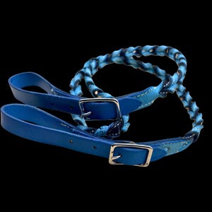 May include: A pair of blue leather stirrup straps with silver-colored buckles. The top portion of the straps are a braided design with blue and black accents. The background is black.