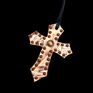 May include: A copper-colored cross pendant with a marbled design and bronze-colored studs. The cross has a small circular emblem at the center and hangs from a black cord. The background is black.
