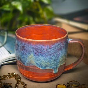 Könnte beinhalten: Ein Keramikbecher mit einer leuchtend orangefarbenen Basis und einer strukturierten, tropfenden Glasur in Blau-, Violett- und Türkistönen, die vom Rand herabfließt. Der Becher hat einen breiten, zylindrischen Körper mit horizontalen Rillen.