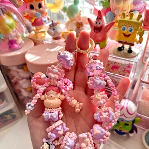May include: A beaded bracelet in shades of pink and purple, featuring small doll-like charms. The bracelet is held in a hand, with a gold-colored clasp. The background includes various figurines and decorative items.