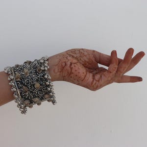 Puede incluir: Un brazalete plateado con detalles intrincados y pequeños adornos redondos. El brazalete se lleva en una muñeca con diseños de henna. El fondo es una superficie blanca lisa.