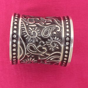 Peut inclure: Un bracelet manchette argenté avec un motif floral et cachemire foncé gravé. Les bords sont bordés d'une rangée de petits points en relief. Le bracelet est posé sur un fond rose.