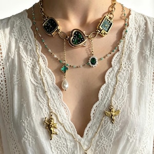 Puede incluir: Un collar dorado de varias capas con colgantes de esfera de reloj, dijes en forma de corazón y adornos colgantes. El collar incluye un trébol de cuatro hojas, una perla y un colgante de cupido. Las esferas del reloj son verdes.
