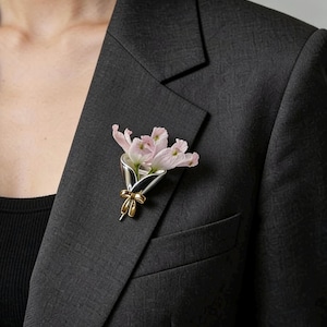 Pode incluir: Broche prateado e dourado em forma de buquê de flores. O broche apresenta um grupo de flores rosa claro e é preso a um blazer cinza escuro. O broche tem um detalhe de laço dourado.