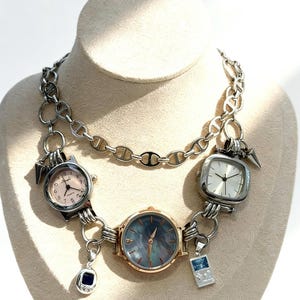 May include: A distinctive necklace with several silver-toned watch faces, showcasing dials in pink and white. The necklace incorporates a silver chain and small charms. The watches are connected to a silver chain.