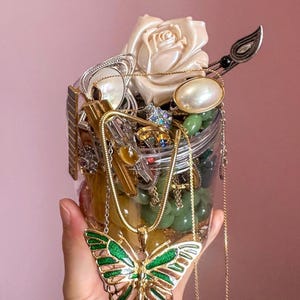 Pode incluir: Um frasco de vidro transparente cheio de várias peças de joalharia, incluindo colares, anéis e brincos. Um pendente de borboleta dourado e verde pende de uma corrente. Uma rosa branca e outros itens decorativos também são visíveis.