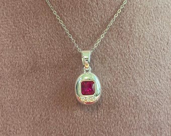 May include: A silver necklace with a pendant featuring a square, deep red gemstone. The pendant is surrounded by a silver frame and small, clear accent stones. The chain is delicate and silver.