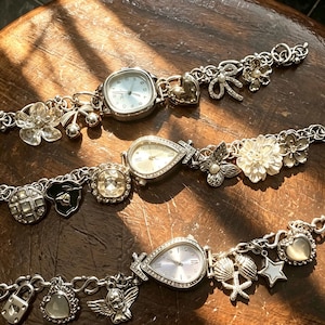 May include: Three silver-tone charm bracelets featuring integrated analogue watches. The bracelets include various charms such as hearts, flowers, butterflies, stars, seashells, a padlock, and an angel figure, all attached to metallic link chains.