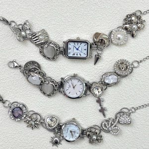 Puede incluir: Tres pulseras de abalorios plateadas con esferas de reloj. Cada pulsera presenta un reloj con esfera blanca y varios abalorios, incluyendo corazones, mariposas y cruces. Las pulseras están conectadas por una cadena y se muestran sobre una superficie blanca.