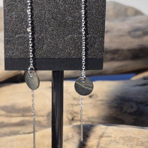 May include: A pair of dangle earrings. Each earring has a small, round, grey stone disc suspended from a delicate silver chain. A silver bar hangs below the stone disc. The earrings are displayed on a black earring holder.