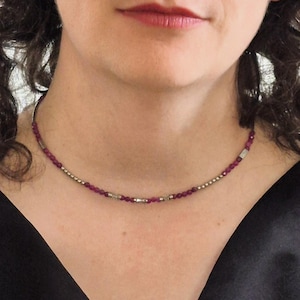 Puede incluir: Un delicado collar con pequeñas cuentas redondas de color rojo intenso intercaladas con detalles plateados. El collar se lleva alrededor del cuello, con una parte superior negra en el fondo.