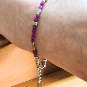 May include: A beaded bracelet featuring small, round, deep pink beads interspersed with silver-colored and gold-colored metal accents. The bracelet has a charm with a small tag hanging from a chain.