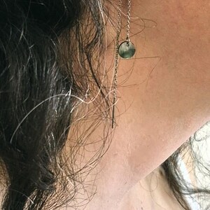 May include: A silver chain earring with a small, round, green-toned charm and a straight bar. The earring is worn in the ear and hangs down. The person's dark hair is visible.