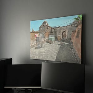 Puede incluir: Una impresión en lienzo de una escena de mapa de videojuego, que presenta un entorno arquitectónico de Oriente Medio con un cielo azul. La obra de arte se muestra encima de dos monitores de ordenador, un teclado y unos auriculares.