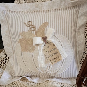 May include: A square throw pillow with a beige and white striped pattern featuring a large white fabric pumpkin appliqué. The pumpkin is decorated with a burlap leaf, a white fabric bow, and a tag that reads In all things Give thanks 1 Thess 5:18.