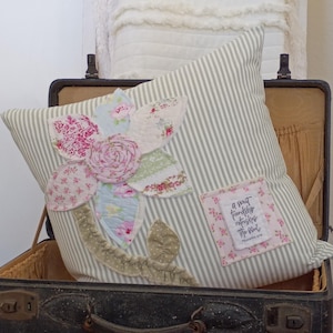 May include: A decorative pillow with a floral design, resting in an open vintage suitcase. The pillow features a striped background and a patchwork flower with pink, blue, and green fabric petals. A small square patch with text is sewn on the pillow.