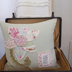 May include: A square throw pillow with a light green and white striped fabric base. It features a large, multi-colored floral applique made of various patterned fabric petals and a central rosette. A small rectangular patch reads: A sweet friendship refreshes the soul.