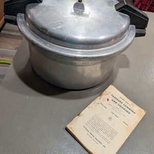 Puede incluir: Una olla a presión de aluminio vintage con asas negras y un manómetro. Un libro usado titulado "Modern Guide to Pressure Canning and Cooking" está al lado de la olla. El libro es de National Presto Industries, Inc.