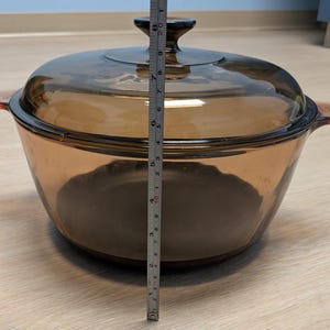 Puede incluir: Una fuente para horno de vidrio transparente de color ámbar con tapa a juego. La fuente tiene dos asas laterales y un asa en la tapa. La altura de la fuente es de aproximadamente 20 cm.