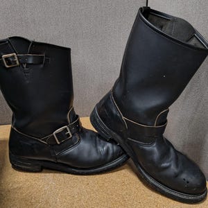 Könnte beinhalten: Ein Paar schwarze Leder-Ingenieurstiefel mit wadenhohem Schaft, abgerundeten Zehenkappen und zwei verstellbaren Riemen mit silberfarbenen Metallschnallen. Ein Riemen befindet sich über dem Spann, der andere am oberen Schaftrand.