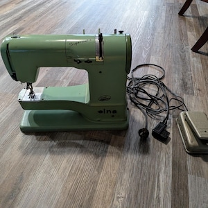 Könnte beinhalten: Eine Vintage Elna Supermatic Nähmaschine in Hellgrün. Die Maschine hat ein Fußpedal und ein Netzkabel. Die Maschine steht auf einem Holzboden.