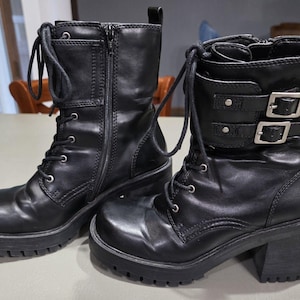 May include: Pair of black leather combat boots with lace-up fronts, side zippers, and chunky soles. One boot features two silver buckles. The boots have a mid-calf height and a block heel.