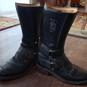 Peut inclure: Une paire de bottes mi-mollet en cuir noir dotées de deux boucles en métal argenté sur le côté de chaque tige. Les bottes sont marquées du logo Chippewa USA, représentant le profil d'une personne portant une coiffe, sur la partie supérieure.