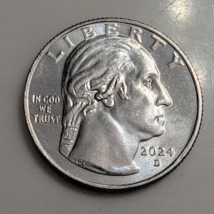 Może przedstawiać: Srebrna moneta ćwierćdolarowa Stanów Zjednoczonych, przedstawiająca profil George'a Washingtona, słowo "LIBERTY" u góry i rok "2024". Moneta zawiera również napis "IN GOD WE TRUST".