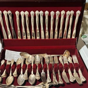 May include: A complete set of silver silverware in a red velvet-lined wooden case. The set includes knives, forks, spoons, and a serving ladle. The silverware has ornate floral detailing on the handles.