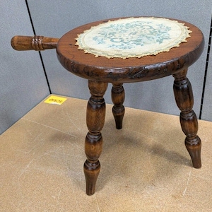 May include: A small, antique wooden stool with a round top and three legs. The stool is stained a dark brown and features a decorative, oval-shaped doily on the seat. A handle extends from the side.
