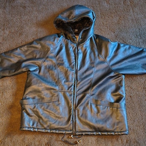 May include: A dark gray hooded jacket with a faux fur-lined hood and cuffs. The jacket has a full-length zipper, two front pockets, and a drawstring at the bottom. The jacket is made of a shiny, leather-like material.