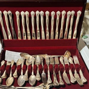 May include: Silver cutlery set in a red velvet-lined wooden case. The set includes knives, forks, spoons, and serving pieces. The handles have a floral design. The case is open, revealing the neatly arranged silverware. A classic dining set.