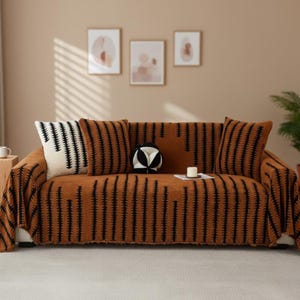 Op de afbeelding: Een bruine bank met een bijpassende sprei en kussens. De sprei en kussens hebben een zwart verticaal streepjespatroon. Decoratieve kussens in zwart en wit liggen ook op de bank. De achtergrond omvat ingelijste kunstwerken en een plant.