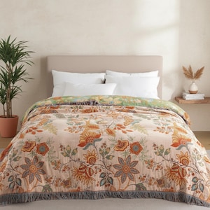 May include: A floral patterned blanket with a peach background and orange, blue, and green accents. The blanket has a fringed edge and is draped over a bed with white pillows. The design features large stylized flowers and foliage.