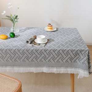 Op de afbeelding: Een grijs en wit patroon tafelkleed met een geometrisch ontwerp. Het tafelkleed heeft een franje en staat op een houten tafel. Een vaas met bloemen, een kop en schotel en een kleine cake staan op de tafel.