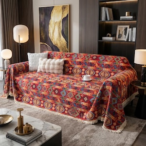 Puede incluir: Una funda de sofá roja con un patrón geométrico en tonos naranja, azul y amarillo. La funda tiene un borde con flecos y está colocada sobre un sofá con cojines blancos. Una taza de café está sobre el sofá.