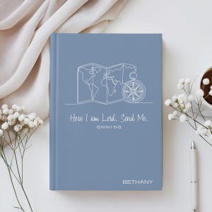 May include: A blue journal featuring a white line drawing of a world map and compass. The cover includes the text "Here I am Lord. Send Me." and "Isaiah 6:8". The name "Bethany" is printed at the bottom of the journal.