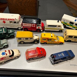 May include: A collection of ten miniature toy ambulances and emergency vehicles, including a yellow ambulance with the text "AMBULANCE" and a red ambulance with the text "AMBULANCE HOPITAUX".