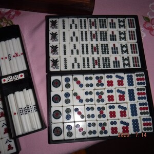 May include: A set of mahjong tiles in a black case. The tiles are white with black and red markings. The tiles are arranged in a grid pattern. The case has a black background with white lettering. The tiles are arranged in a grid pattern. The case has a black background with white lettering. The tiles are arranged in a grid pattern. The case has a black background with white lettering.