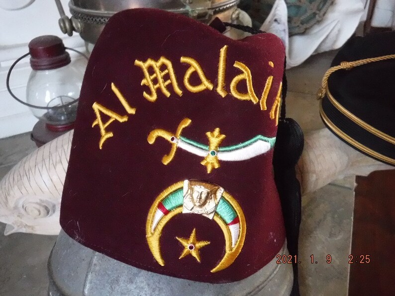 May include: A maroon fez with gold embroidery that reads "Al Malai" and features a crescent moon, star, and crossed swords.