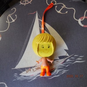 May include: A yellow plastic doll with blonde hair and orange clothing. The doll is hanging from a string and has a smiling face.
