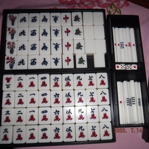 May include: A set of mahjong tiles in a black carrying case. The tiles are white with black and red characters and symbols. The case has a black interior and a black exterior with a silver trim.