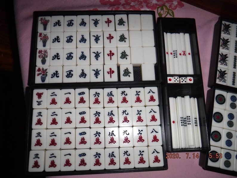 May include: A set of mahjong tiles in a black carrying case. The tiles are white with black and red characters and symbols. The case has a black interior and a black exterior with a silver trim.