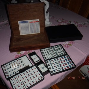 May include: A wooden box with a black lid containing a set of mahjong tiles. The tiles are arranged in a grid pattern and are made of white plastic with black and red markings. The box is open and the tiles are visible. The box is sitting on a pink tablecloth with a floral pattern.