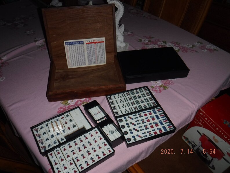 May include: A wooden box with a black lid containing a set of mahjong tiles. The tiles are arranged in a grid pattern and are made of white plastic with black and red markings. The box is open and the tiles are visible. The box is sitting on a pink tablecloth with a floral pattern.