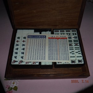 May include: A wooden box containing a set of white mahjong tiles with black characters and red accents. The box also includes two instruction cards with Chinese text and a set of white sticks. The box is closed.