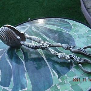 May include: Silver-toned serving tongs shaped like seahorses with a shell-shaped handle. The tongs are on a green and blue mosaic surface.