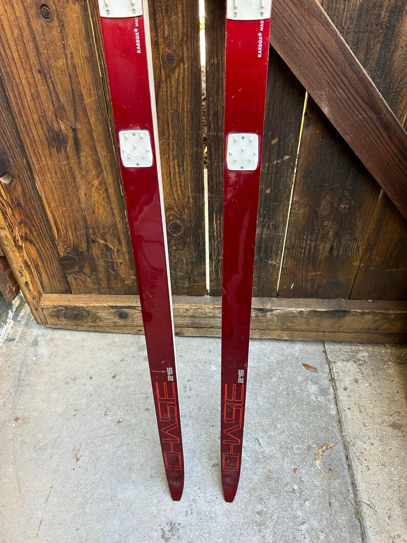 May include: A pair of red cross-country skis with black bindings and the word "CHASE" printed in white on each ski. The skis are 215 cm long.