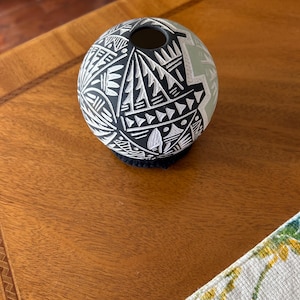 May include: A spherical black and white ceramic vase with a geometric pattern. The vase has a small opening at the top and sits on a dark, textured base. The vase is on a wooden table.